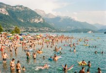 Canicule en Franche-Comté : où se baigner en toute sécurité cet été ? Many people swimming and relaxing at a crowded lakeside beach with mountains in the background