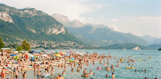 Canicule en Franche-Comté : où se baigner en toute sécurité cet été ? Many people swimming and relaxing at a crowded lakeside beach with mountains in the background