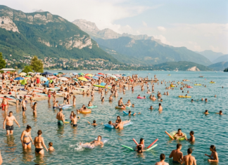 Canicule en Franche-Comté : où se baigner en toute sécurité cet été ? Many people swimming and relaxing at a crowded lakeside beach with mountains in the background