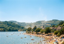 Canicule en Bourgogne : où se baigner en toute sécurité cet été ? Crowded lakeside beach with swimmers, sunbathers, umbrellas, and small boats near forested hills